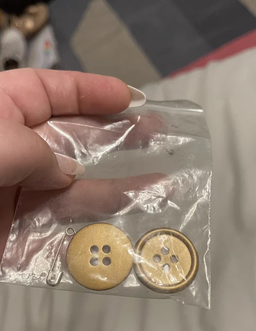 Picture of a hand holding a small, clear plastic bag containing two round, gold buttons.