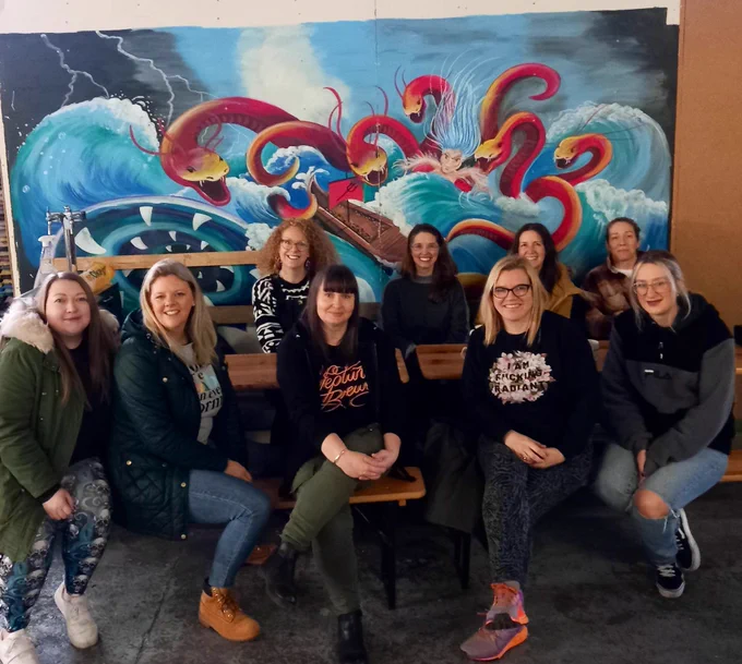 A happy group of women from posing on benches at Neptune’s new brewery in front of the sea monsters mural