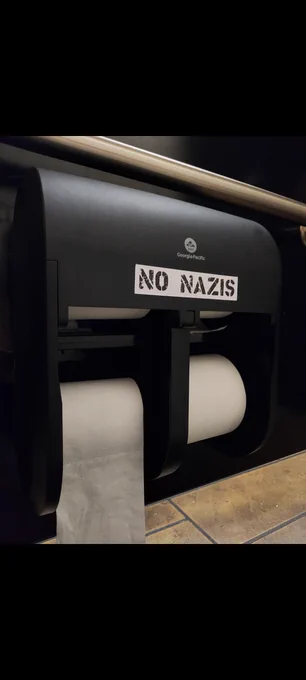 A public toilet paper dispenser labeled "NO NAZIS."