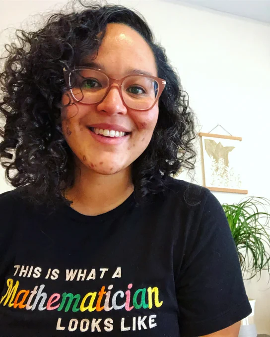A Black woman wearing glasses and a black shirt with the words "This is what a Mathematician looks like" written on it.