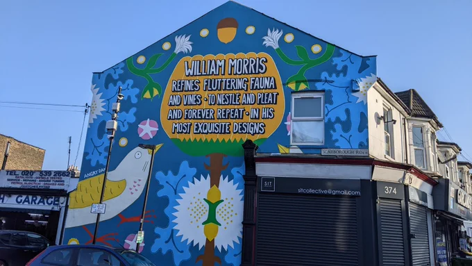 Mural on a wall on the end of a row of houses and shops. The image is a blue background with a yellow and white bird with flowers in white and yellow with an acorn and a tree on the middle. The tree has the words 'William Morris refines fluttering fauna and vines to nestle and pleat and forever repeat in his most exquisite designs' in the centre.