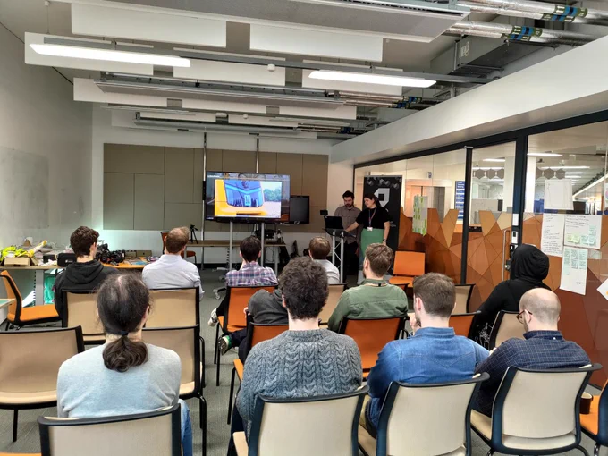 Daniel and Emily stand at the front doing their presentation. There is the audience in front of them. On the screen is a video of the Tyne and Wear Metro. 