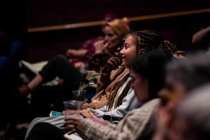 The row of an audience in a cinema auditorium. The person in the middle of the row's face is illuminated as they smile at the stage.