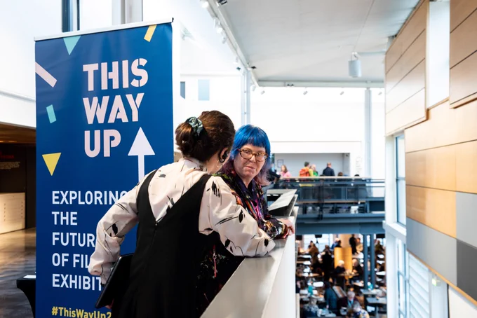 Two people stand leaning against a balcony wall in Dundee Contemporary Arts, with a dark blue This Way Up banner behind them.