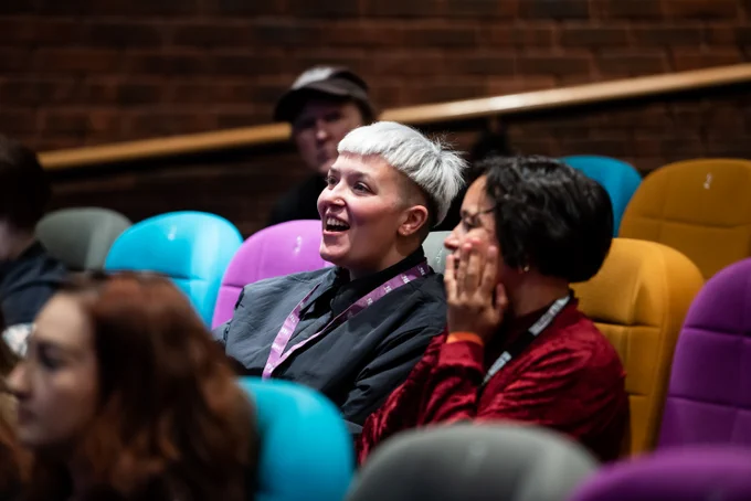 Two people sit in a cinema on colourful seats talking to each other. One is wearing a black shirt with blonde hair and the other is wearing a red jumper with short dark hair.