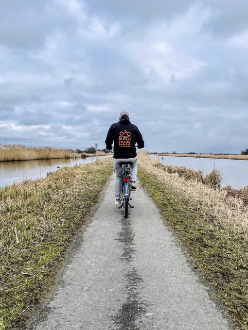 A man in a grey jeans and a black hooded jacket rides a bicycle on a grey asphalt path in a nature area in South Holland. He is surrounded on both sides by water.