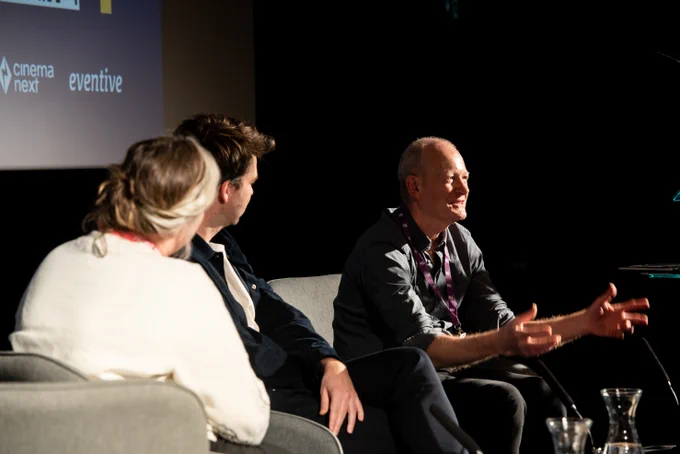 Three people sit on a stage facing an audience - the one on the far right is speaking directly to them and gesturing with their hands.