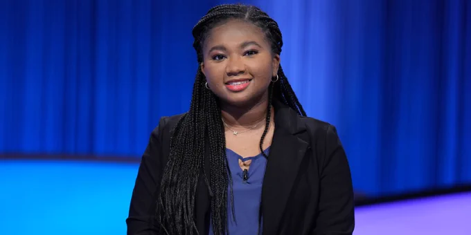 Emory senior Maya Wright stands behind her lectern on the Jeopardy! stage dressed in a blue top and black jacket