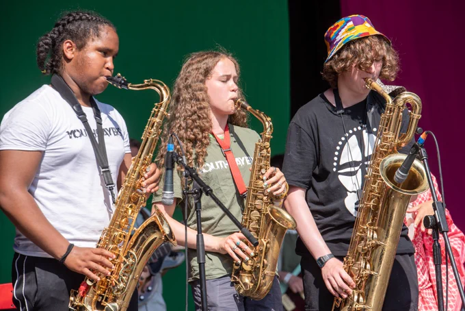 Image is of a band called Youthsayers performing on the Village Green stage in 2022. It includes 3 young people playing saxophone.