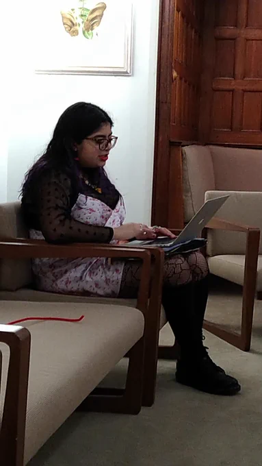 Eulalia reading from her laptop, seated in black boots,  black top, and white flowery dress 