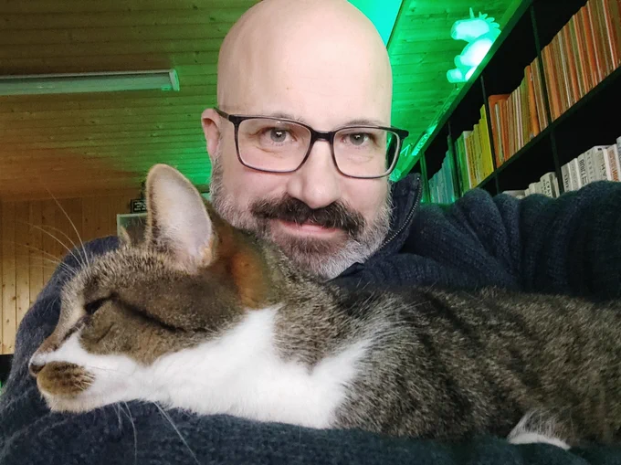 Stephen, a smiling bald man with a grey beard holding and a sleepy cat