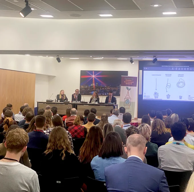 Image of audience facing panel members who are sat on the left hand side of the room. Large screen of the right displaying powerpoint presentation