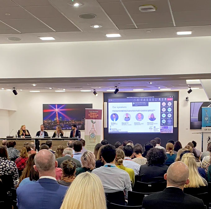 Image of audience facing panel members who are sat on the left hand side of the room. Large screen of the right displaying powerpoint presentation