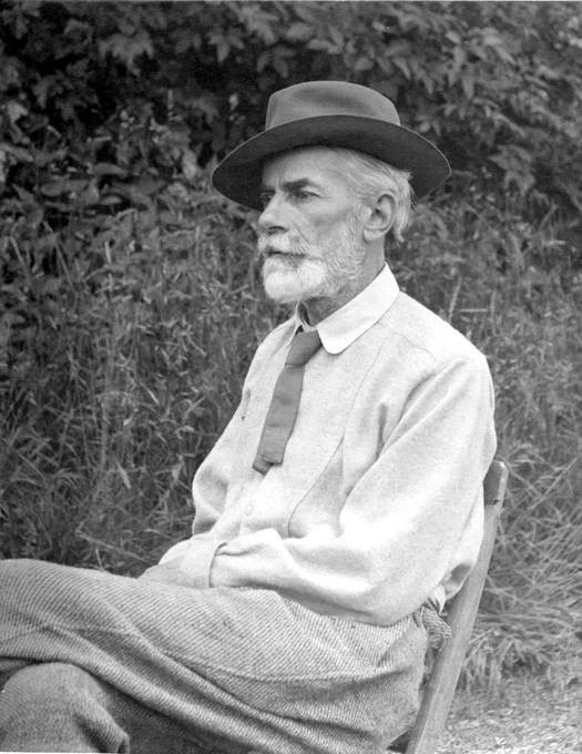 Edward Carpenter sitting on chair in a garden. He has grey hair and a beard, and is wearing a hat, a shirt with a tie, and corduroy trousers.