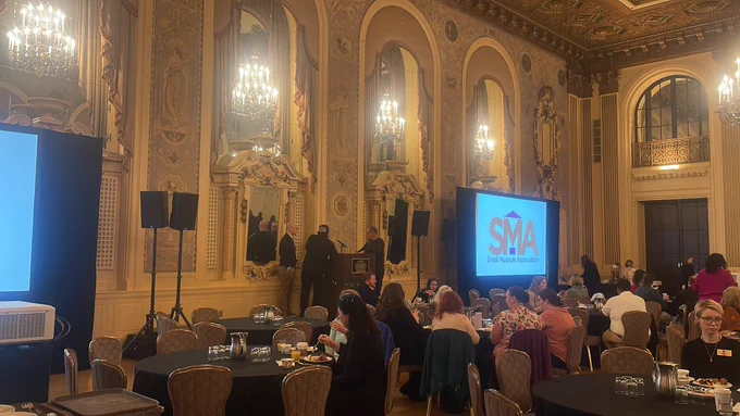 Full ballroom with a podium and two screens with the SMA logo. People sitting and eating at tables.