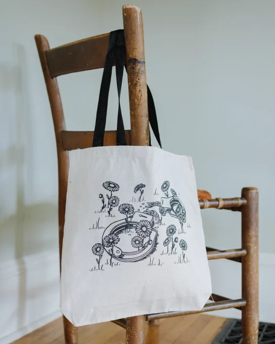 photo of a cream colored tote bag with black handles hung on a wooden chair. in the center of the bag is a design of handcuffs, laying broken on the ground surrounded by blooming flowers.