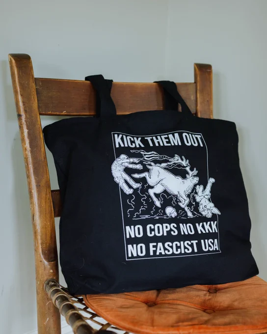 a photo of a black tote bag sitting on a wooden chair. white design on the bag says "kick them out" and shows an elk kicking a cop and running over a klanmember. bottom text on the bag reads "no cops no kkk no fascist usa"
