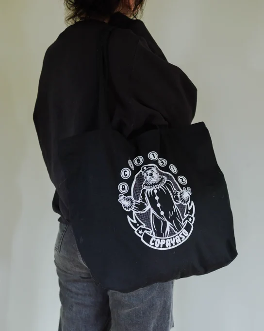 a person facing away from the camera wearing a black tote bag, black shirt and black jeans. tote bag has a white illustration of a clown on it, surrounding the clown are letters and numbers "ACAB" and "1312," inside juggling balls. below the clown, a banner of text reads "copayaso." artist: ProtestPapi