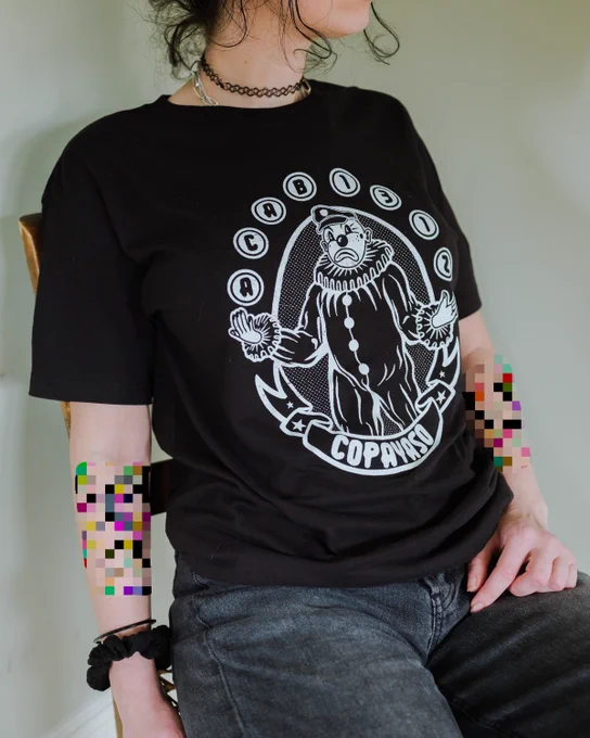 photo of a person sitting down, wearing a black t shirt with an illustration of a white clown. ink on the shirt is white. surrounding the clown are numbers and letters reading "ACAB" and "1312" in juggling balls. below the clown a banner reads "copayaso." artist: ProtestPapi