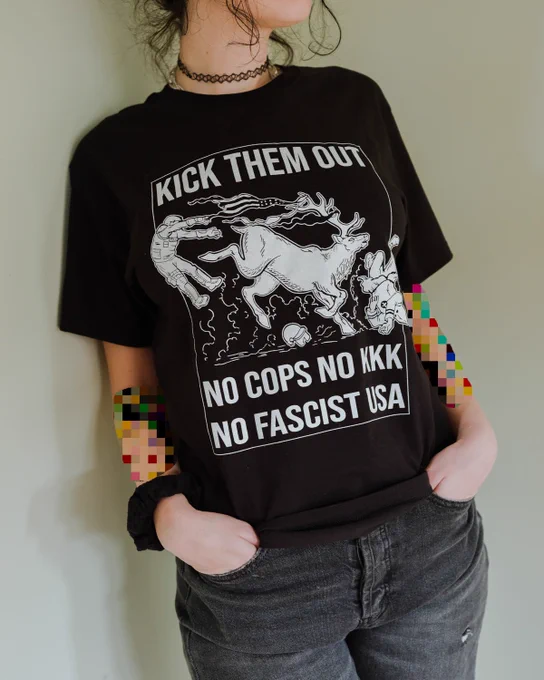 a photo of a person wearing a black shirt sleeved shirt, on the shirt there's a white ink illustration of an elk kicking a riot cop, and running over a klanmember. text above the elk reads "kick them out" and below the elk reads "no cops no kkk no fascist usa."
