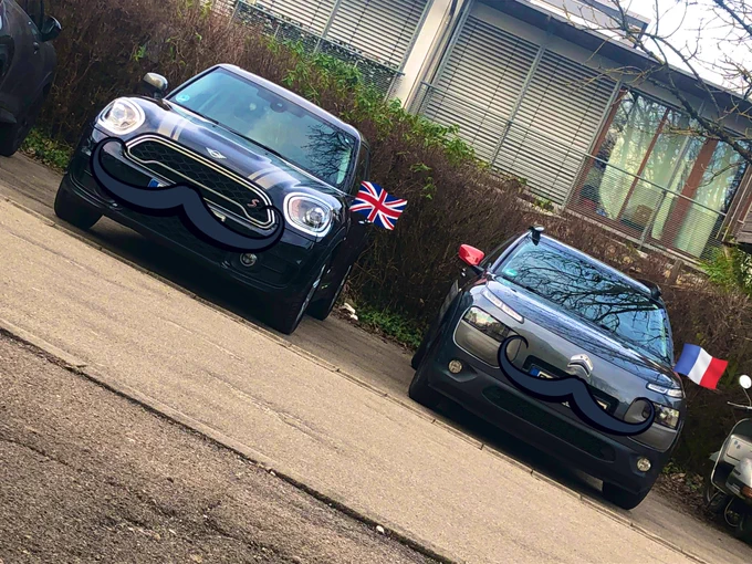 Perlas Autos nebeneinander. Links ein schwarzer MINI Countryman, rechts ein grauer Citroën C4 Cactus mit roten Außenspiegeln.
Um die Kennzeichen zu verdecken, hat Perla mit den Twitter Bildbearbeitungsprogramm jeweils einen Schnurrbart eingefügt und passend zu den Autos die entsprechende Länderflagge über dem rechten Außenspiegel.