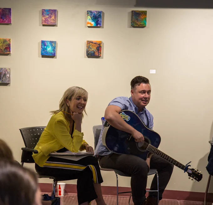 Taes Leavitt and Nick Adams sit in chairs engaging with students. Nick holds a blue acoustic guitar in his lap.