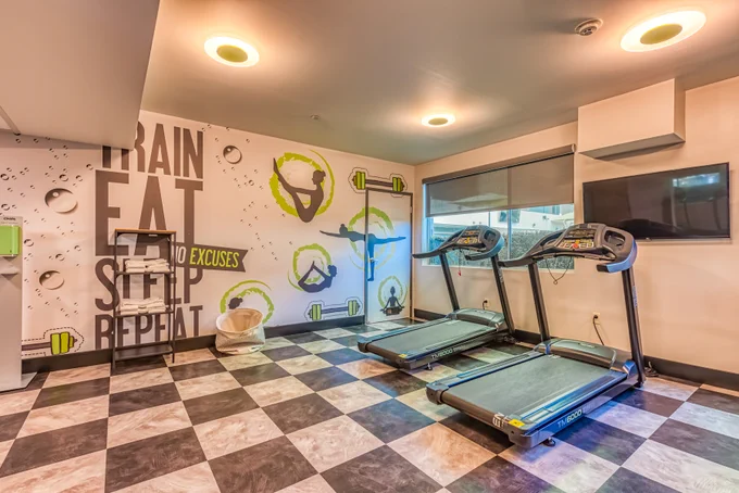 Lexen North Hollywood Fitness Center: 2 Treadmills, TV, Water station. Checkered flooring and wall depicting images of different exercises and text: Train, Eat, Sleep, Repeat No Excuses.