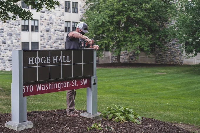 A person wearing a baseball cap drills together a sign with the words "Hoge Hall" in front of the grey Hokie Stone Hoge Hall. 