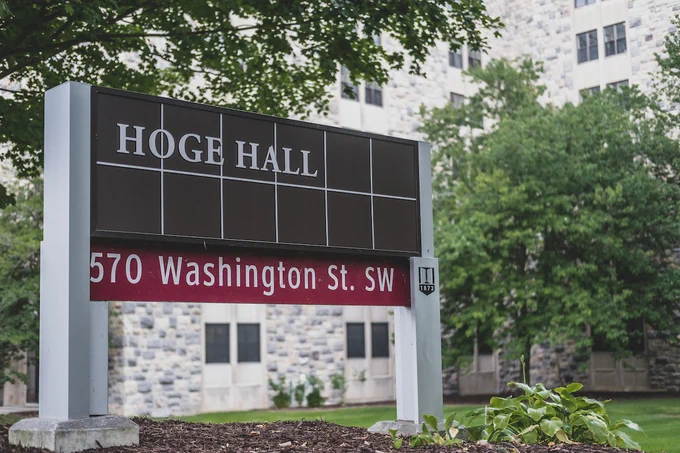 A sign with the words "Hoge Hall" is pictured on a grass lawn in front of the grey Hokie Stone Hoge Hall. 