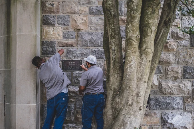Two people wearing grey shirts remove a plaque reading "Lee Hall" on a grey Hokie Stone building. 