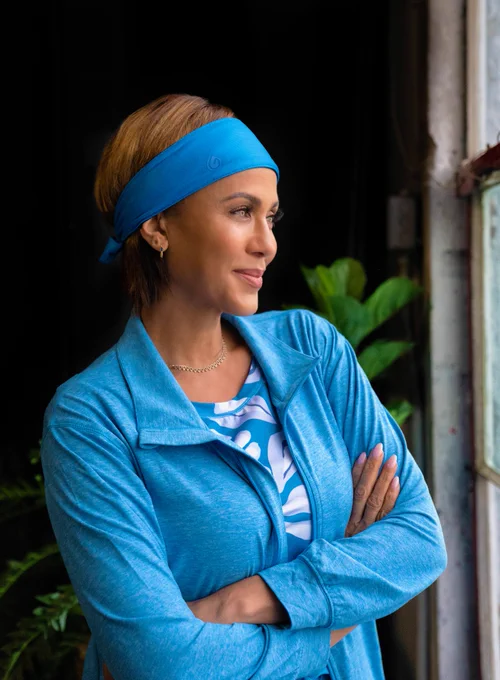 Photo of Gymwrap creator Nicole Ari Parker smiling with her arms folded across her body, staring out a window. She has medium-light brown skin, and short brown hair with golden highlights. She’s wearing a light blue headband, light blue long sleeved zip-up jacket and purple/blue floral activewear tank top, all from Gymwrap.