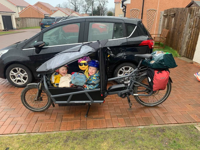 A parked cargo bike on a rainy drive. Three kids in the box, a big parcel and a pannier on the rack