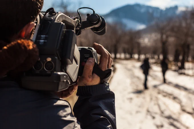 Man holding a film camera outside pointed at a road with two figures walking a long it. Mountains can be seen in the distance.