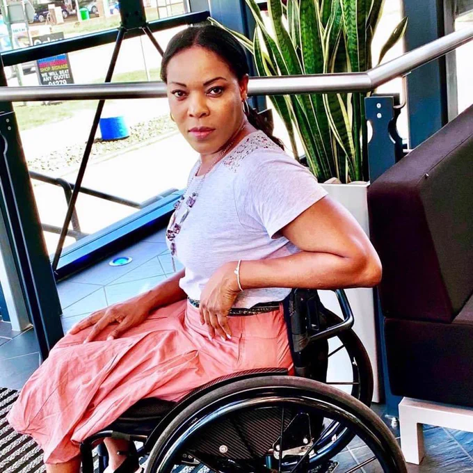A lady sitting in a wheelchair. Wearing a pink skirt and a grey short sleeve top. Her leg elbow is resting on her wheelchair handle.