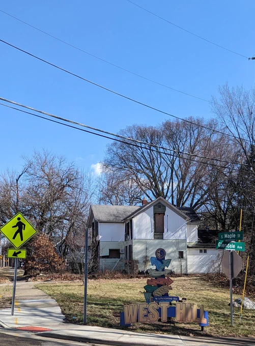 Picture in Akron Ohio at S Maple St and Glendale Ave. Shows art sign that says "Welcome To West Hill". Behind it is a neglected abandoned house that looks like it caught on fire, house is all boarded up and pile of trash bags around it