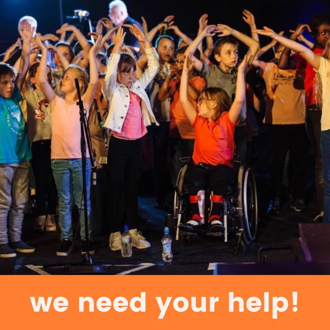 A big group of children raise their hands as they sing on stage. One of the children is in a wheelchair. This is part of our annual schools' singing events, sing out and sing   sign.