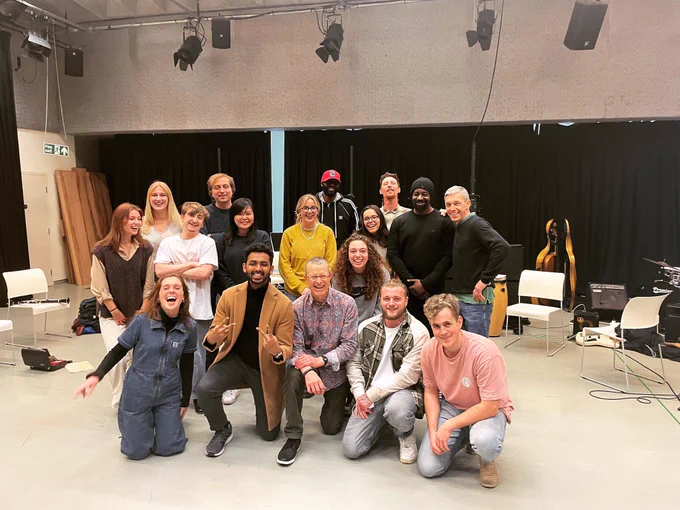 The photo shows a group of happy people posing together in a big room with musical instruments and equipment in the background