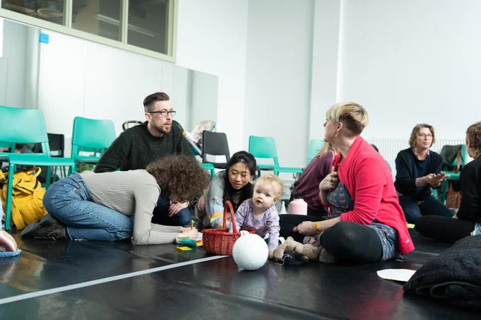 A group of artists with a young baby taking part in an artist development workshop. The group is discussing a task with materials including a basket, ball, pens, pencils and paper. 
