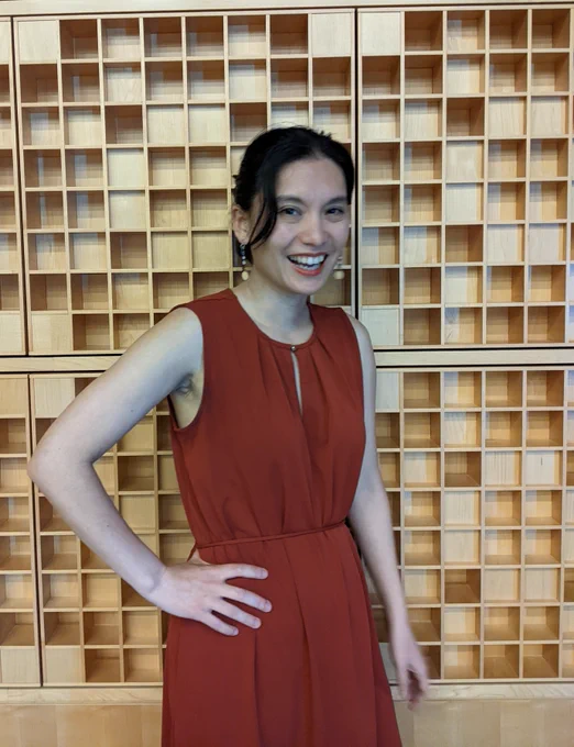 Regina Barber wearing a dress, is smiling. She stands in front of a wall of small wooden boxes, with her hand on her hip.