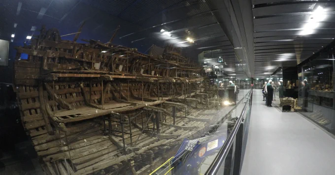 A view of a wooden shipwreck, with people walking along a glass-walled gallery along side it