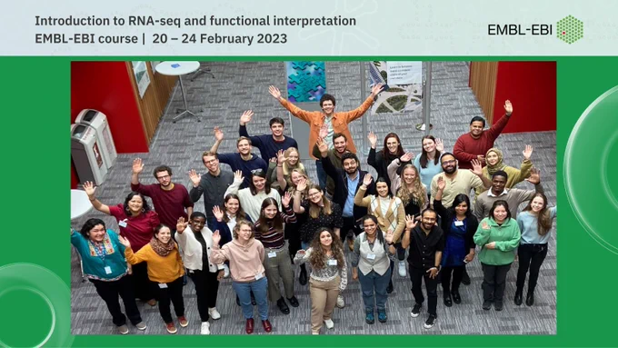 A group of smiling people are looking up at a camera and waving. You can see scientific posters and red walls behind them. The overlaid text says: Introduction to RNA-seq and functional interpretation. EMBL-EBI course |  20 – 24 February 2023. The EMBL-EBI logo is also displayed. Photo credit: Sophie Spencer, EMBL-EBI, 2023.