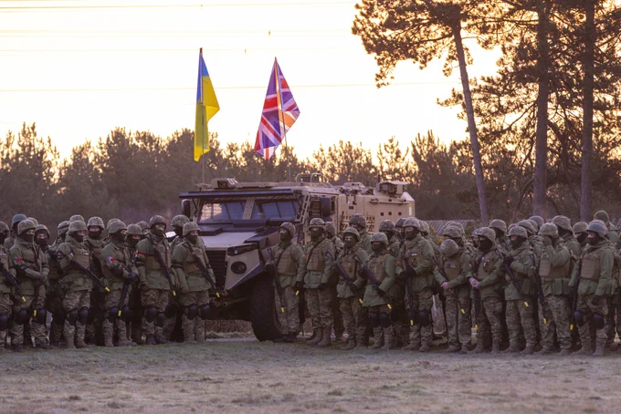 A group of Ukrainian troops stand in front of an armoured vehicle holding the Ukrainian and UK flags.