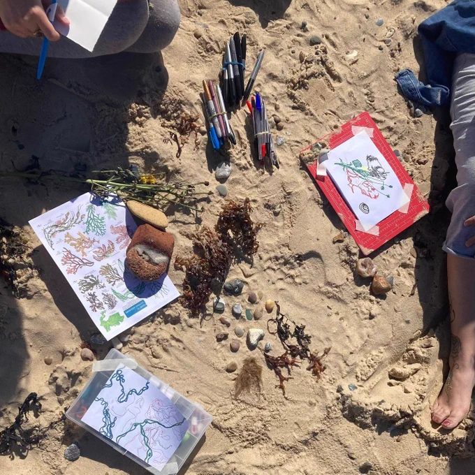  looking down onto a sandy beach dotted with collected pebbles and seaweed, with pens and art materials. Taped on boards are two drawings of the collected materials and two people's legs can be seen at the top left and right edge of the image, as they're taking part in the drawing