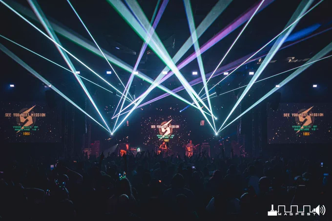 On display is a shot from Super MAGFest Melee, namely Metroid Metal performing live on the main stage towards the end of their set. A panoply of blue-white, blue, and purple lasers criss cross over those metal virtuosos, and behind them and mirrored on two screens to either side is a Metroid style banner proclaiming that the crowd's rate of rocking is 100%. The foreground is full of people, eagerly watching Metroid Metal tear it up on stage.