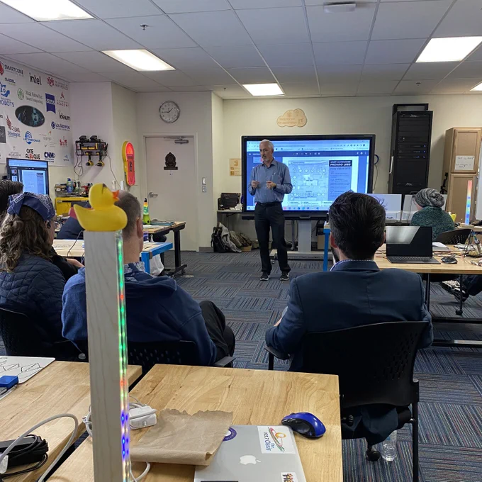 Brian Osterloh, Department of Technology and Innovation Director, speaks to a group of people in an IoT Bootcamp classroom. In the foreground are desks with laptops and various chargers. In the background is a digital display board with a page from the Smart City presentation featuring an overhead map of the Pino Yards Proving Grounds with icons for all of the technology that's being tested there, like smart lights.