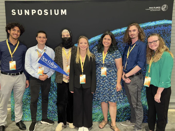 Seven people standing behind the ‘Sunposium’ backdrop, one professor and six students, smiling at the camera and holding a New College flag