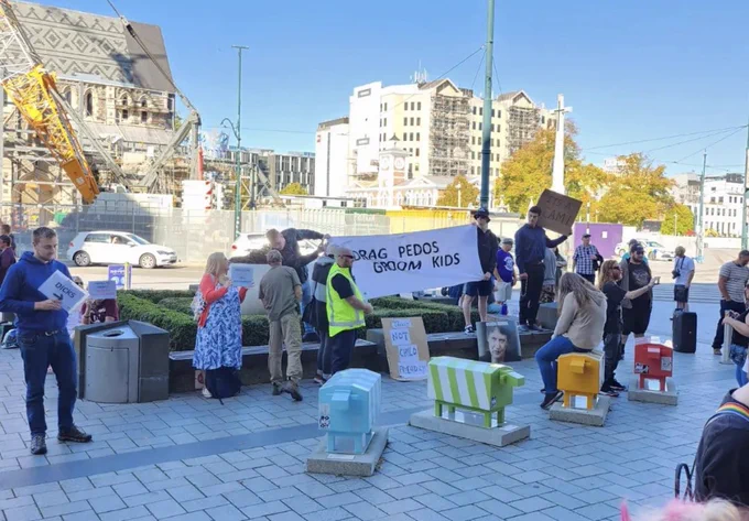 Small group of people holding signs claiming that drag queens and LGBTQIA  people are groomers and pedophiles. A large banner reads: drag queens groom kids. There are approximately 12-14 men pictured, and 2-3 women, all middle aged   and white.