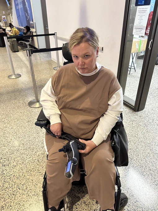 Jessica a white woman sits in her power wheelchair at the airport with the wheelchair joystick broken in her lap