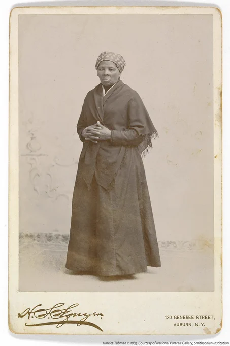Black and white photo of Harriet Tubman standing wearing  a dark ensemble consisting of a shawl and a long-sleeve, floor length dress. She has a patterned scarf tied over her hair and her hands are folded and rested on her midriff. There is a signature for the photographer, H. Seymour Squyer in the bottom left side corner of and printed text that reads "130 GENESEE STREET, AUBURN, N.Y."  in the bottom right corner. 