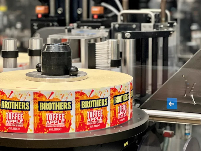 A photograph of a thick roll of labels, still stuck to the backing paper, wrapped around a piece of machinery. The labels feature the red silhouette of an apple with an orange splash design printed over the top. On top of these designs is the Brothers logo, followed by white text reading, 'toffee apple English cider'. 